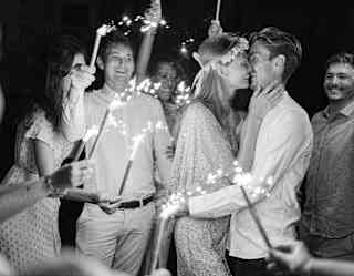 A couple embraces and smiles surrounded by friends holding sparklers at night. The scene is joyful and celebratory, with everyone dressed in light-coloured clothes. The photo is in black and white.