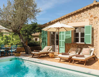 Elegant sun loungers with floral cushions face the private pool of a charming villa with mint green shutters and blue bistro set