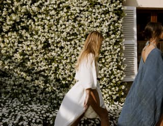 A female guest with long blond hair, wearing a white linen wrap dress strides past the jasmine-clad hotel, seen side on.