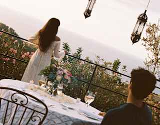 Angled image of a man at a dining table as he gazes at a woman in a white dress with long dark hair, standing on the terrace.