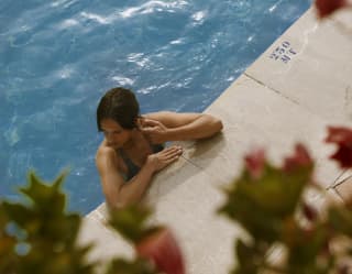 A dark-haired woman in the sparkling blue pool relaxes at the edge with her elbows resting on the stone, seen from above.