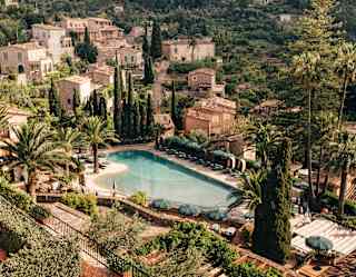 Stunning view over the hotel's gardens, tropical-shaped pool and stone residences, with palms and pines, seen from above.