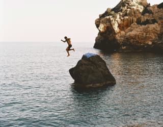 Sun catches on a limestone headland of Cala Deià where a man leaps from a large rock into the shadowy water, taken mid-air.