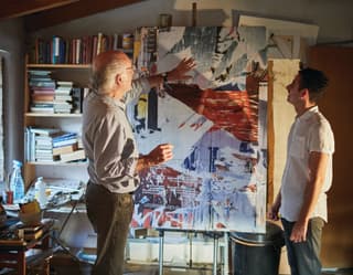 Artist in a studio describing a modern art painting on an easel to a guest