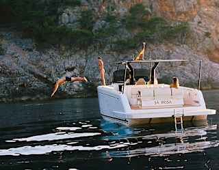 A man is captured mid-air diving from the Sa Brisa boat, moored in a bay, as a female friend watches from the deck.