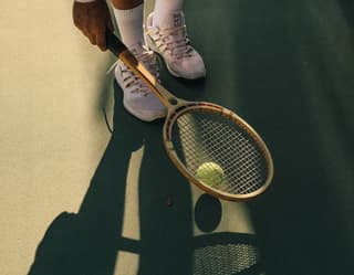 A man in tennis whites uses an old wooden racquet to bounce and scoop up a yellow ball. A low sun casts long shadows