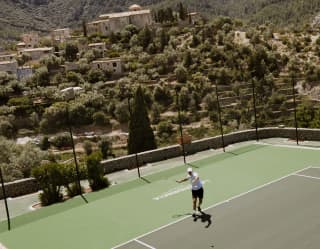 A player swings his racket arm on a GreenSet tennis court with Sant Joan de Deià church on the hill behind, seen from above.