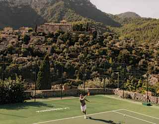 A man hits a tennis ball from the baseline, in a view of one end of a green court, with a craggy forested hillside backdrop.