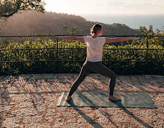 A woman in a white top and black leggings adopts the yoga warrior pose in gardens facing the sea, seen from behind.