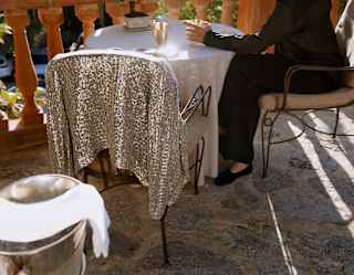 Seen behind a chill bucket, a sequined jacket rests on a chair at a terrace table, where a guest in black drinks Champagne.