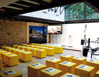 Rows of lemon yellow cubes offer bench seating in an airy venue space with a glass atrium room and exposed stone walls