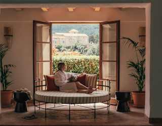 A woman in a robe relaxes on a day bed, gazing out at sunny garden views through open doors, seen across the indoor spa pool.