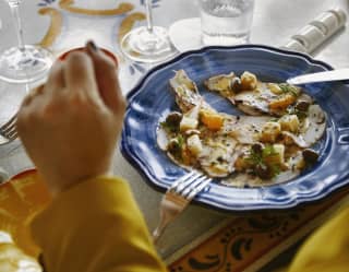 A guest rests her hands on the table by her lunch of focaccia, grilled vegetables and olives, served on a blue china plate.