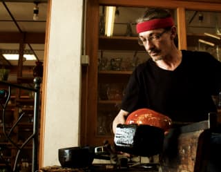 Glassblower Nicolas Diverchy, in a black top and red headband, makes a glass orange bowl in his Verrerie du Marais workshop.