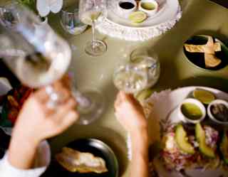 A hand holds a glass of white wine at a table of diners enjoying fresh tacos with vibrant toppings, guacamole and bean dip.