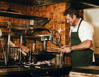 A man wearing a white shirt and green apron grills food in a restaurant kitchen, focusing on the food while using tongs. The background features brown tiled walls and stainless steel kitchen equipment.