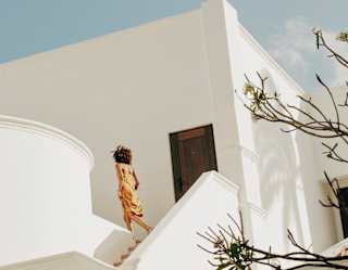 A woman in a soft orange dress enters a white residence from an external staircase, viewed from gardens below.