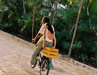 A woman rides on the back of a bike, holding onto a yellow box behind her as the male rider sets off along the stone path.