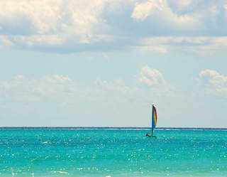 On glittering turquoise sea, a Hobie Cat Catamaran with red, yellow and blue sail, heads towards the dark blue horizon.
