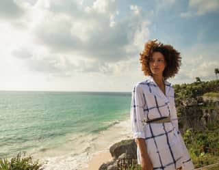 A lady in a blue and white shirt and breeze in her dark curly hair, stands at a viewpoint in front of the Riviera Maya coast.