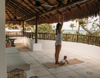 A woman stretches up as she practices yoga on a mat with her baby, on a private terrace with woven lanterns and sea views.