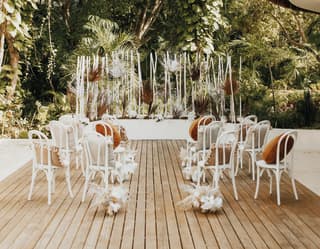 On a wooden deck surrounded by jungle, a registry table and witness chairs with white flourishes await a memorable ceremony.