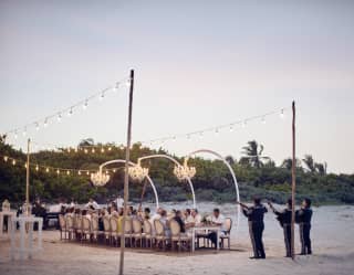 Guests at a banquet table on the beach at sunset, lit by fairy lights