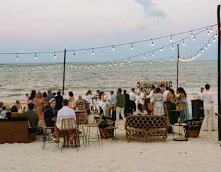 A celebration party mingles on an area of beach in front of the ocean equipped with seating, bar and suspended lights.