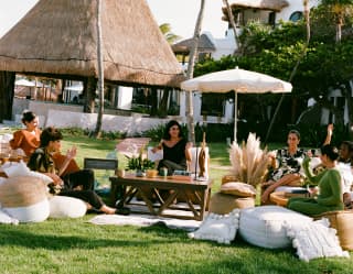 With a backdrop of palms and a palapa, five guests lounge on sofas and cushions, enjoying a private event in Coronas Garden.