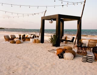 Pescados Beach is ready for an event with cushions and chairs on the sand either side of a pergola, and strings of lights.