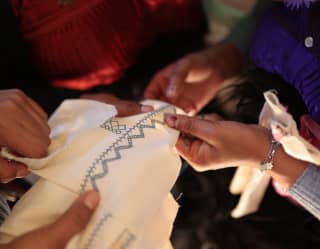 An embroiderer stitches track and zigzag patterns on to white cotton as another craftsperson holds it taut, seen in closeup.