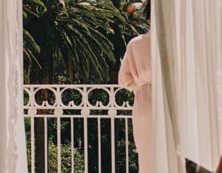 A woman in a pink robe leans on the white railings of her balcony, gazing over lush palm trees, seen from behind a curtain.