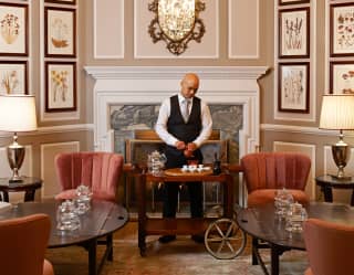 A male staff member in a black waistcoat prepares hot drinks on a trolley in an opulent and warmly-furnished lounge area.