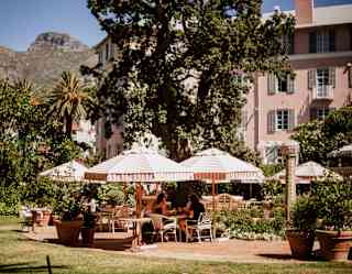 Four guests dine in the shade of large coral and white-striped parasols on a flower-filled terrace, with a mountain backdrop.