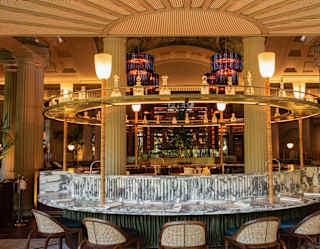 A circular grey marble counter, circled by chairs, orbits a glowing Art Deco-style circle bar with a ring of decanters above.