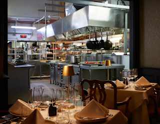 View from the Chef's Table dining room over two laid tables and into the spotless kitchen where a tray of produce is visible.