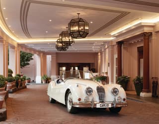 A vintage white Jaguar XK140 car waits in the paved, colonnaded entrance to the hotel beneath twinkling lights and lanterns.