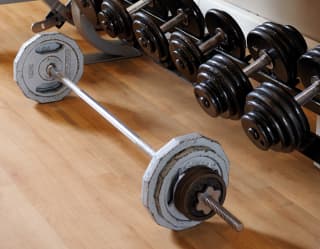 Fitness centre detail of a weights rack with rows of black dumbbells and a long silver weighted barbell on the floor.