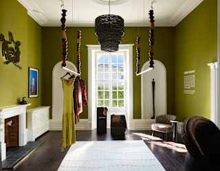 A well-lit room with olive green walls, a large window, hanging wooden rails displaying clothes, a fireplace, wicker furniture, and a basket-like chandelier. Sunlight streams through the window onto a white rug.