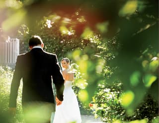 Seen through tree branches, a bride in white looks back at her groom in a dark suit as they stroll in the sunny garden.