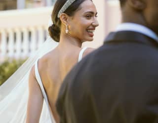 Stepping ahead of the groom, a bride in a low-backed dress, with tied-back dark hair, tiara and veil looks back with a smile.