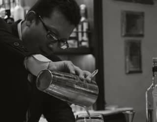 A barman decants a freshly-shaken cocktail from a silver shaker into a glass at the marble bar, shot in black and white.