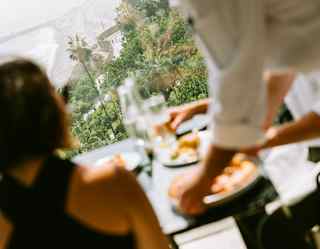 Angled image of a chef placing plates on a table where two guests dine overlooking the Lima coast, in soft-focus.