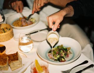 View over a table at Tragaluz, as a man pours cream sauce onto his vibrant main, with bread, water and Negroni-style drink.