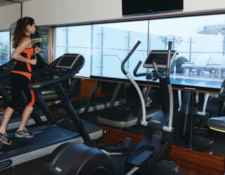 A female guest enjoys using equipment in the hotel gym and fitness room with the pool through the window