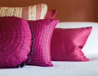 Multiple shaped cushions in cerise, cream and russet lie on a white bed framed by theatrical style curtains