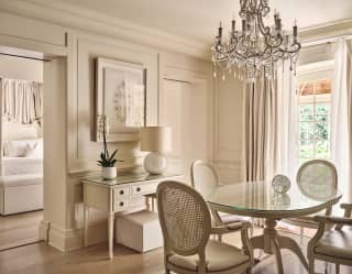 A crystal chandelier hangs above a glass-topped table with four white chairs in a dining area with pale furniture and décor.