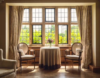 Two French dining chairs and a side table, with a cloth and flower vase, sit in bay window looking on to luminous foliage.