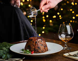 Brandy drizzled from a tiny pan coats a Christmas pudding with holly sprig decoration as festive fairy lights twinkle behind.