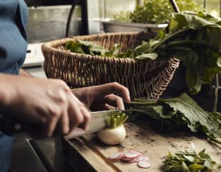 A chef cuts a small white turnip in half. On the chopping board are slivers of radish and a wicker basket overflowing with leaves